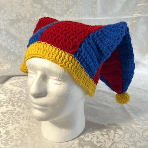 May include: A handmade crochet jester hat in red, blue, and yellow. The hat features a red top, blue side panels, and a yellow brim and pom-poms. The hat is displayed on a white mannequin head.