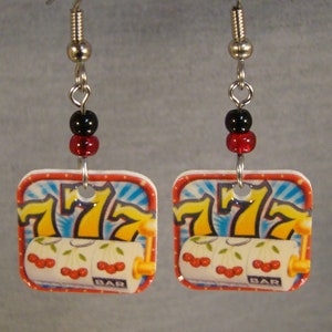 May include: A pair of square earrings featuring a slot machine design. Each earring has a red and black bead above a square pendant with a casino-themed graphic. The graphic includes the number 7, cherries, and the word "BAR".