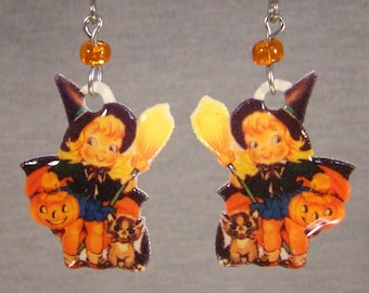 May include: A pair of Halloween-themed earrings. Each earring features a vintage illustration of a smiling child witch holding a broom and a jack-o'-lantern. The earrings have orange beads and silver-tone ear wires.