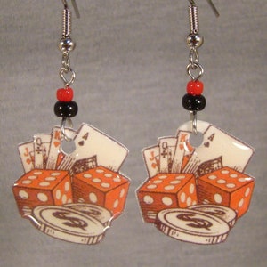 May include: Dangling earrings with a gambling theme. Each earring features a design of orange dice, playing cards, and coins. The earrings are suspended from silver-tone hooks with red and black beads.