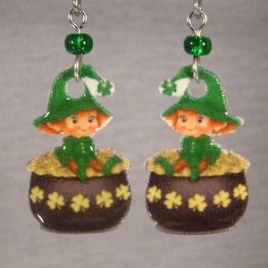 May include: A pair of St. Patrick's Day earrings. Each earring features a cartoon leprechaun wearing a green hat with a white shamrock, sitting in a pot of gold. The pots are dark brown with yellow shamrock designs.