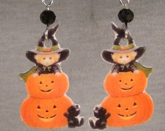 May include: A pair of Halloween-themed dangle earrings. Each earring features a smiling orange pumpkin with a witch and a black cat. The earrings have silver-colored hooks and a black bead.