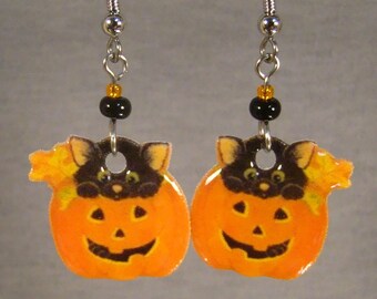 May include: A pair of Halloween-themed earrings. Each earring features a smiling orange pumpkin with a black cat peeking out. The earrings are suspended from silver-colored hooks with black and gold beads.