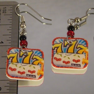 May include: A pair of square earrings featuring a slot machine design. The earrings have a red border, a white background with cherries, and the number 7. The earrings are approximately 1 inch in length.