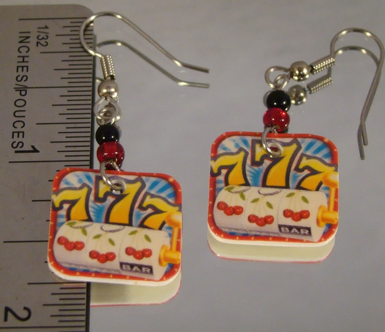 May include: A pair of square earrings featuring a slot machine design. The earrings have a red border, a white background with cherries, and the number 7. The earrings are approximately 1 inch in length.