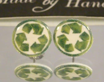 May include: Handmade stud earrings featuring a white background with a green recycling symbol. The earrings have a green glittery border. The text "Made by Hand" is visible in the background.