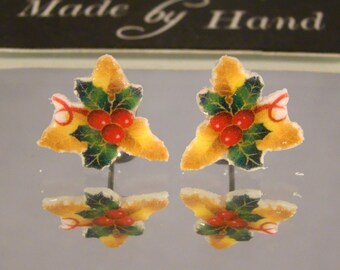 May include: A pair of holly leaf earrings with red berries and green leaves. The earrings are yellow and white with a frosted appearance. The text "Made by Hand" is visible in the background.