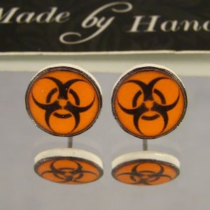 May include: A pair of orange and black stud earrings featuring a biohazard symbol. The earrings have a silver post and a white border. The text "Made by Hand" is visible in the background.