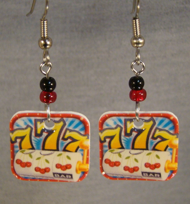 May include: A pair of square earrings featuring a slot machine design. Each earring has a red and black bead above a square pendant with a casino-themed graphic. The graphic includes the number 7, cherries, and the word "BAR".