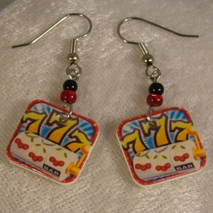 May include: A pair of dangle earrings featuring a slot machine design. Each earring has a square charm with a casino-themed graphic, including the number 777, cherries, and the word "BAR". The earrings are accented with black and red beads.