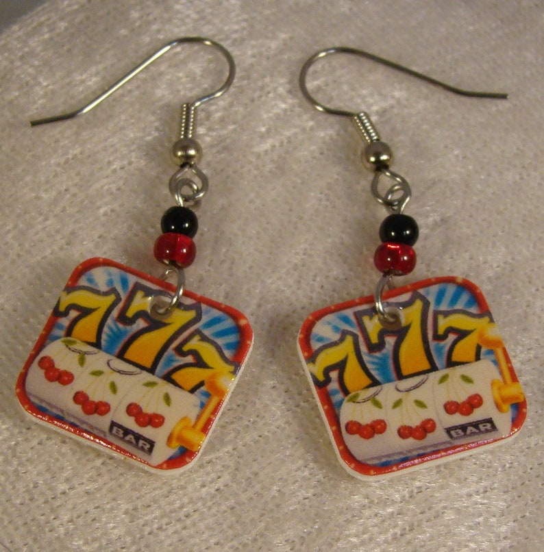 May include: A pair of dangle earrings featuring a slot machine design. Each earring has a square charm with a casino-themed graphic, including the number 777, cherries, and the word "BAR". The earrings are accented with black and red beads.
