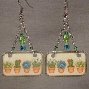 May include: A pair of dangle earrings featuring rectangular pendants with a botanical design. Each pendant displays a row of potted succulents and cacti. The earrings are accented with green and blue beads and silver-tone chains.