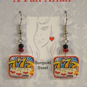 May include: Unique handmade earrings featuring a slot machine design. The earrings have a square shape with a red border, displaying the numbers 777, cherries, and the word "BAR". The earrings are made with surgical steel and have black and red beads.