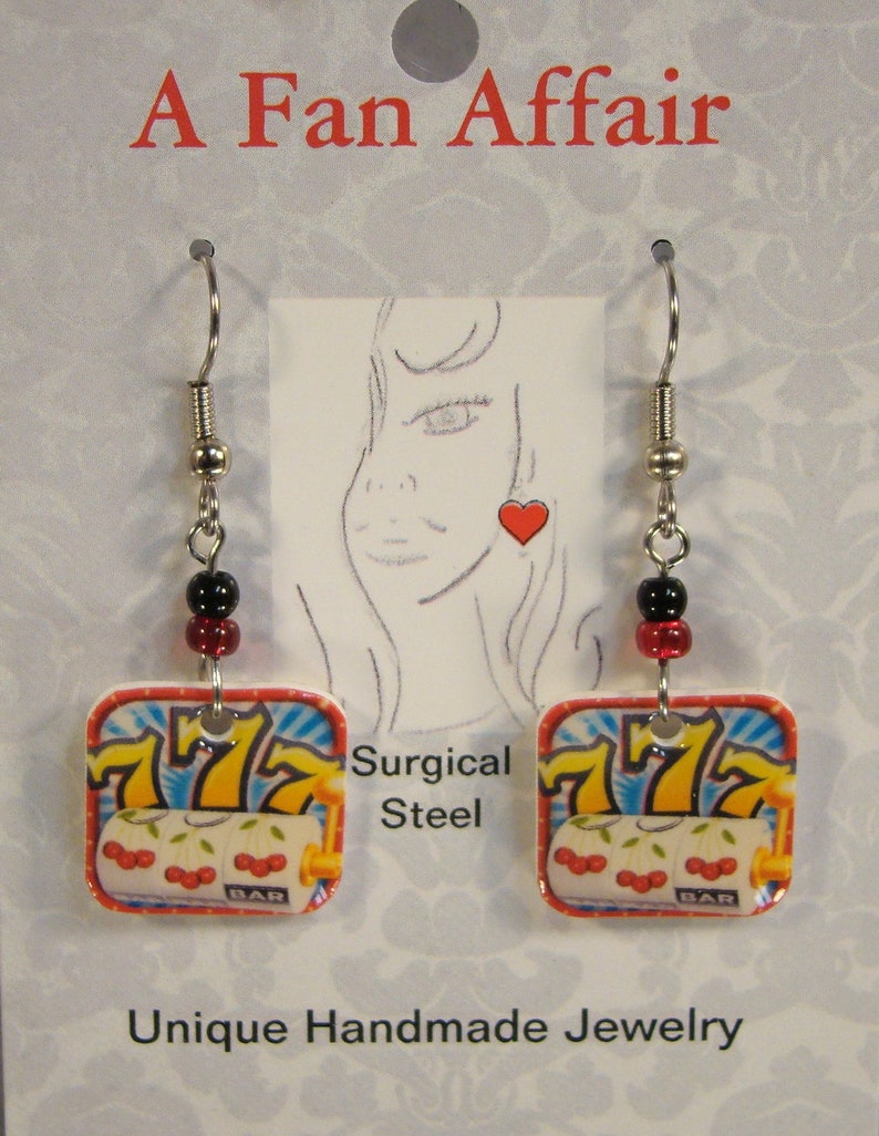 May include: Unique handmade earrings featuring a slot machine design. The earrings have a square shape with a red border, displaying the numbers 777, cherries, and the word "BAR". The earrings are made with surgical steel and have black and red beads.