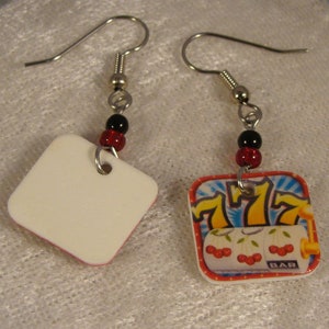 May include: A pair of dangle earrings. One earring features a white square with a red border, the other has a slot machine design with cherries, sevens, and the word "BAR". Each earring has a silver hook and is accented with black and red beads.