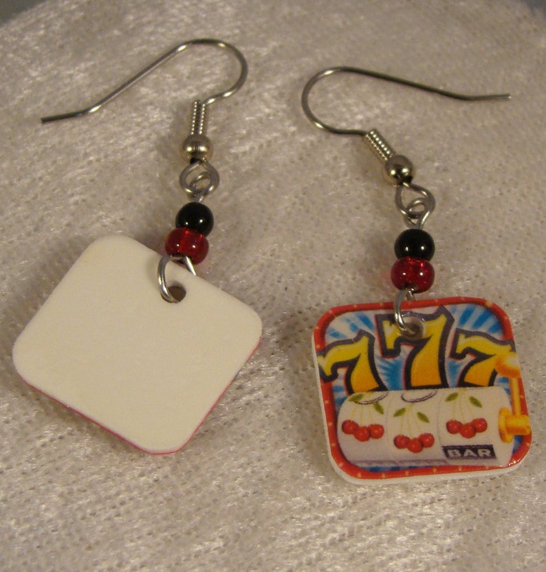 May include: A pair of dangle earrings. One earring features a white square with a red border, the other has a slot machine design with cherries, sevens, and the word "BAR". Each earring has a silver hook and is accented with black and red beads.