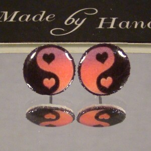 May include: Pair of round stud earrings featuring a yin-yang design with heart accents. The earrings have a black and orange color scheme, with the text "Made by Hand" in the background.