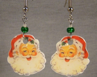 May include: A pair of Christmas-themed earrings featuring Santa Claus faces. Each earring has a red and white Santa hat, a smiling face, and a white beard. The earrings are connected with silver-colored hooks, a green bead, and a small white bead.