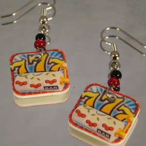 May include: A pair of square-shaped earrings featuring a slot machine design. The earrings have a white border and are adorned with red and black beads. The slot machine graphic displays cherries, the number 7, and the word "BAR".