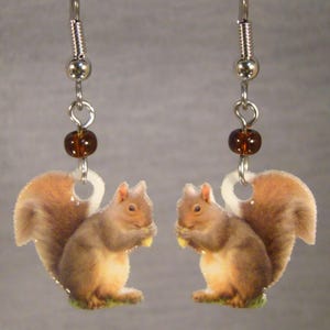 May include: Squirrel earrings with a detailed illustration of a squirrel holding a nut. The earrings have a silver-colored hook and a brown bead. The squirrel's fur is brown and tan, with a white tail.