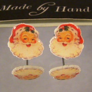 May include: Two Christmas-themed earrings featuring Santa Claus faces with red hats and white beards. The earrings are attached to a small, round base with a Santa Claus image. The text "Made by Hand" is visible at the top.