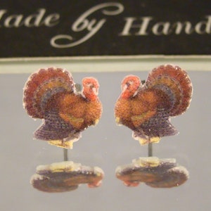 May include: A pair of stud earrings featuring a detailed graphic of a turkey with brown, orange, and red plumage. The earrings are positioned in front of a black sign with the white text Made by Hand. The turkey design is mirrored on each stud.