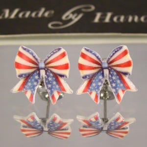 May include: A pair of bow-shaped stud earrings with a patriotic design. The bows feature red and white stripes with a blue section adorned with white stars, set on silver posts. The text "Made by Hand" is visible in the background.