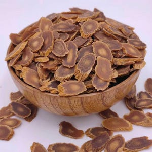 Könnte beinhalten: Eine Holzschale gefüllt mit geschnittenen, getrockneten Ginsengwurzeln. Die Scheiben sind hellbraun mit einem dunkleren braunen Inneren. Die Schale steht auf einer weißen Oberfläche. Einige Ginsengscheiben sind verstreut.