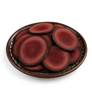 May include: A collection of small, round, burgundy-colored ceramic plates with dark brown edges are arranged in a decorative, brown, woven-style bowl. The plates have a speckled texture and a glossy finish, creating a rustic aesthetic.