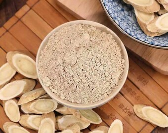 May include: A bowl of light brown powder is the focal point, with sliced beige root pieces scattered around it. The bowl rests on a wooden surface, and a patterned plate with more root slices is visible in the background.