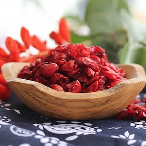 May include: A wooden bowl filled with dried, bright red goji berries. The bowl has a natural wood grain and sits on a patterned blue and white cloth. Red flowers are visible in the background.