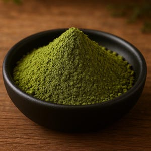 以下が含まれることがあります： 鮮やかな緑色の抹茶パウダーが入った黒いボウルのクローズアップ。きめ細かく粉末状になったパウダーがボウルの中央に高く積み上げられています。ボウルは木製の表面に置かれており、質感と色のコントラストを生み出しています。