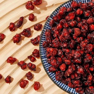 Puede incluir: Primer plano de arándanos rojos secos, algunos esparcidos sobre una superficie de madera clara, otros apilados en un cuenco de cerámica con dibujos azules y blancos. Los arándanos son de un color rojo intenso.