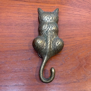 May include: A brass cat-shaped hook, viewed from the back, with detailed fur texture. The hook is in a sitting position with its tail curled. The metal has a warm, golden hue, and the hook is mounted on a wooden surface.