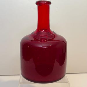 May include: A deep red glass bottle with a rounded base and a long, narrow neck. The bottle has a smooth, shiny surface and is likely used for decorative purposes or as a vase. The glass is a rich, dark red colour.