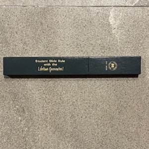 Puede incluir: Una caja rectangular verde oscuro con letras doradas que dice "Student Slide Rule with the Lifetime Guarantee!". La caja también tiene un logotipo circular dorado y el número "341 3526".