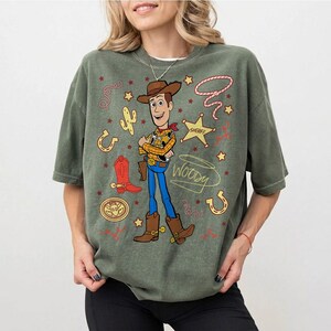 May include: Olive green t-shirt featuring a cartoon illustration of Woody from Toy Story. Woody is depicted in his cowboy attire, surrounded by themed graphics like a sheriff's badge, cactus, and horseshoe. The word "Woody" is written below the character.