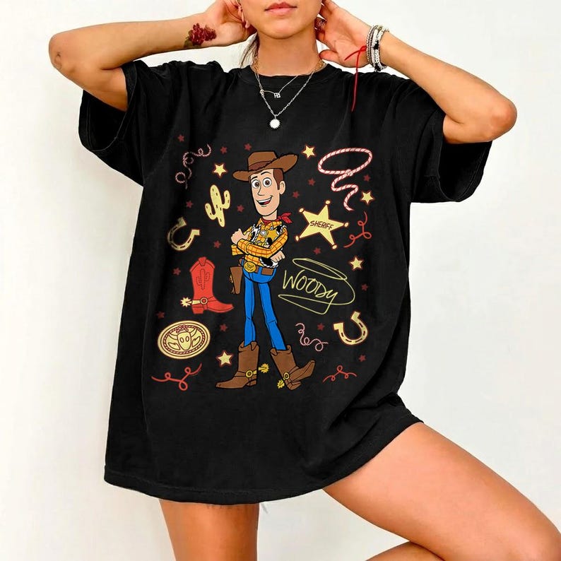 May include: Black t-shirt featuring a cartoon illustration of Woody from Toy Story. The design includes Woody in a cowboy hat, boots, and sheriff's badge, along with the word "Woody" in a playful font. Additional graphics include a cactus, horseshoe, and stars.