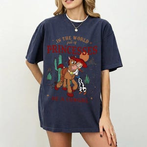 以下が含まれることがあります： カウガールと馬の漫画のグラフィックが特徴のネイビーブルーのTシャツ。テキストは「IN THE WORLD full of PRINCESSES BE A COWGIRL」です。デザインにはサボテンと星が含まれています。