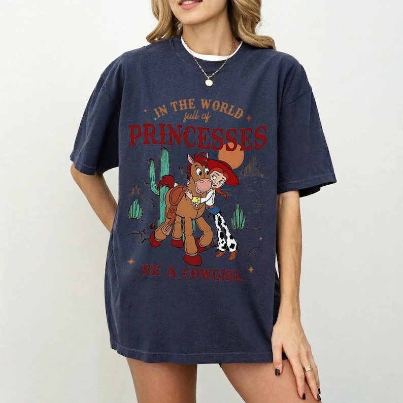 以下が含まれることがあります： カウガールと馬の漫画のグラフィックが特徴のネイビーブルーのTシャツ。テキストは「IN THE WORLD full of PRINCESSES BE A COWGIRL」です。デザインにはサボテンと星が含まれています。