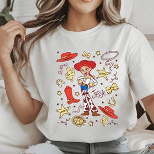 Peut inclure: T-shirt blanc avec une illustration de dessin anim&eacute; de Jessie de Toy Story. Le motif comprend un chapeau rouge, des bottes, un cactus, une &eacute;toile de sh&eacute;rif et le nom "Jessie". Le t-shirt a un col rond et des manches courtes.