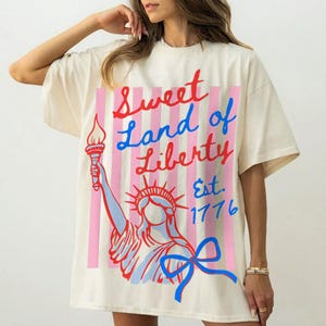 May include: Cream-colored oversized t-shirt featuring a graphic of the Statue of Liberty with the text "Sweet Land of Liberty Est. 1776". The design includes red, blue, and pink accents.