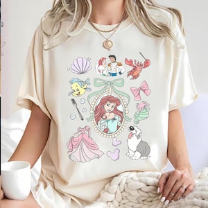 May include: A cream-coloured t-shirt with various cartoon illustrations from a popular animated film. The design includes characters such as Ariel, Flounder, Sebastian, and Prince Eric, along with other related elements.