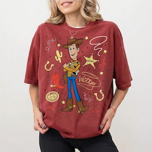 May include: A maroon t-shirt featuring a cartoon image of Woody from Toy Story. Woody is depicted in his cowboy attire, with a sheriff's star, horseshoe, and cactus graphics. The word "Woody" is written in a stylized font.