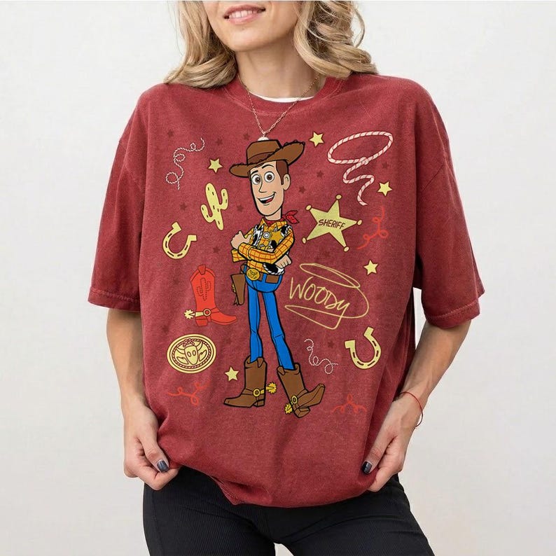 May include: A maroon t-shirt featuring a cartoon image of Woody from Toy Story. Woody is depicted in his cowboy attire, with a sheriff's star, horseshoe, and cactus graphics. The word "Woody" is written in a stylized font.