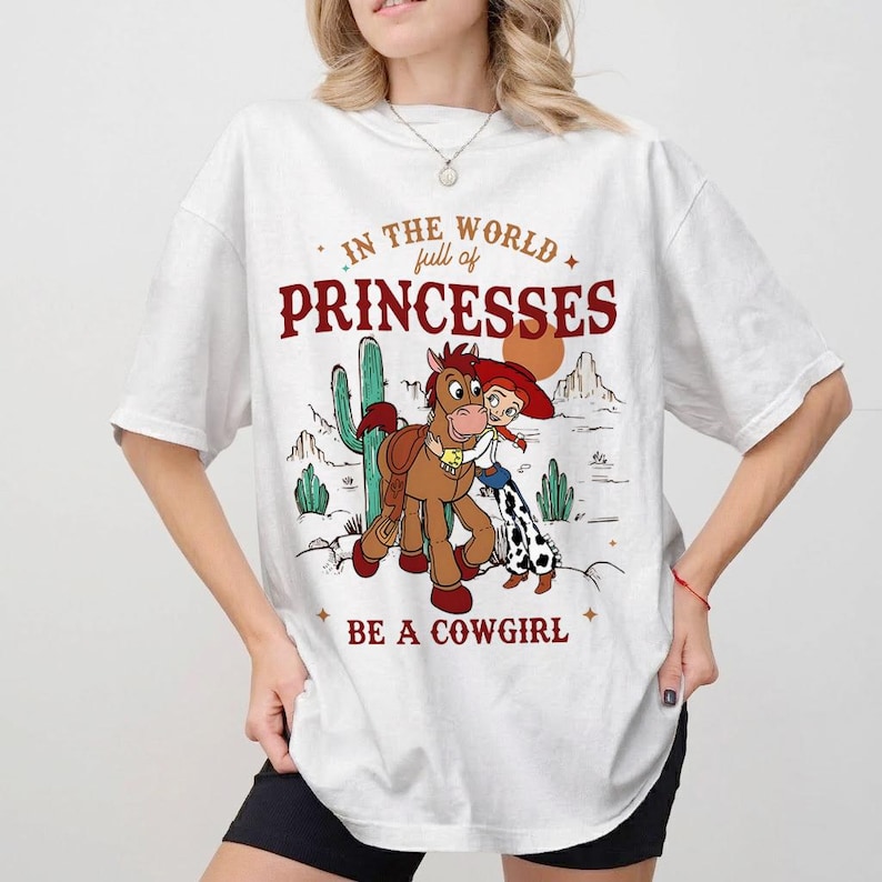 以下が含まれることがあります： 漫画の馬、カウガール、そして「IN THE WORLD full of PRINCESSES BE A COWGIRL」のテキストが特徴の白いTシャツ。 デザインには砂漠の風景要素と星が含まれています。