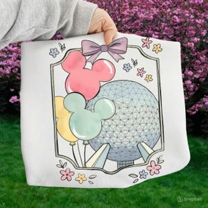May include: White tote bag featuring a colorful design with Mickey Mouse balloons, a light blue geometric sphere, and a purple bow. The design is framed with a black border and floral accents. The bag is held by a person.