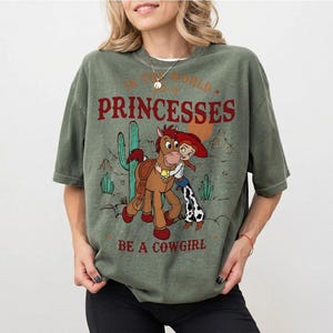 以下が含まれることがあります： オリーブグリーンのTシャツ。漫画の馬とカウガールが描かれ、「IN THE WORLD of PRINCESSES BE A COWGIRL」の文字が入っています。デザインにはサボテンと砂漠の風景が含まれています。