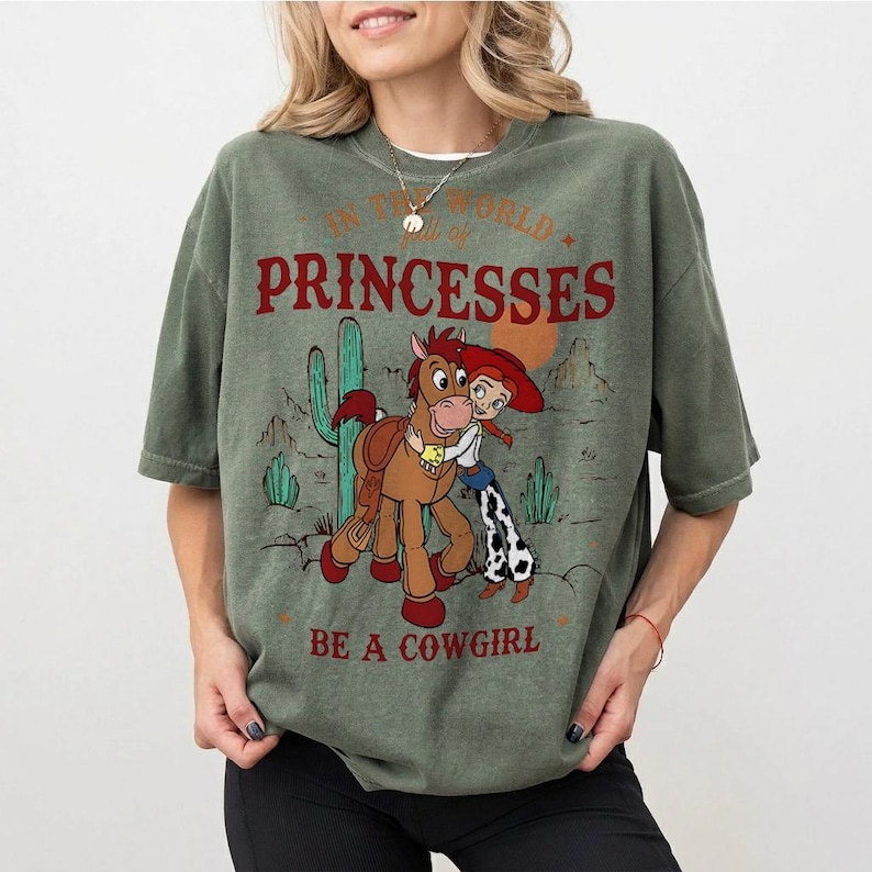 以下が含まれることがあります： オリーブグリーンのTシャツ。漫画の馬とカウガールが描かれ、「IN THE WORLD of PRINCESSES BE A COWGIRL」の文字が入っています。デザインにはサボテンと砂漠の風景が含まれています。