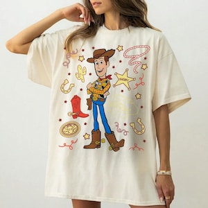 May include: Cream-colored t-shirt featuring a cartoon illustration of Woody from Toy Story. The design includes Woody in his cowboy attire, surrounded by western-themed graphics like stars, a sheriff badge, and a horseshoe. The shirt is a casual, oversized fit.